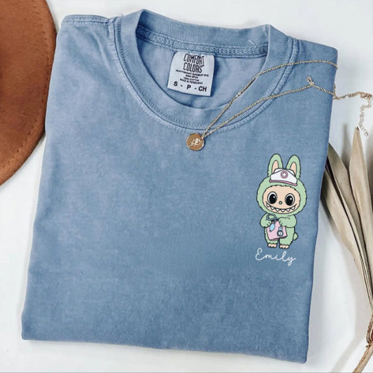 Folded blue t-shirt with a cartoon character and 'Emily' text, next to a brown hat and dried leaves.